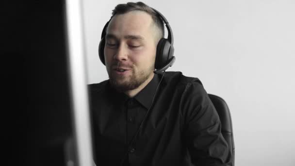 Young businessman or student in a shirt sitting against monitor of computer in office with a white background in headphones and speaking with somebody via internet with a happy face. Bussines call. — Stock Video