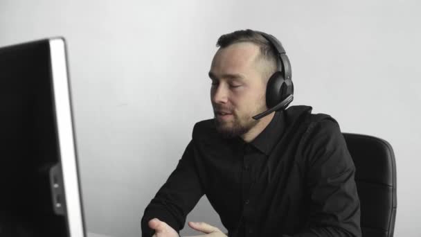 Joven hombre de negocios o estudiante en una camisa sentada contra monitor de computadora en la oficina con un fondo blanco en los auriculares y hablando con alguien a través de Internet con una cara feliz. Llamada de negocios . — Vídeos de Stock