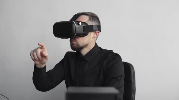 Joven barbudo sentado contra la computadora con gafas de realidad virtual tratando de tocar algo o tocar algo delante de lo que ve. Tecnologías modernas. El concepto de tecnología futura . — Vídeos de Stock