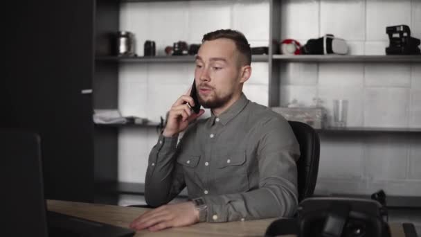 Jonge zakenman of student in een shirt zittend tegen een laptop en met zijn zakenpartner pratend per smartphone. Werken aan een pc aan een tafel in het kantoor met een doordachte uitdrukking. — Stockvideo