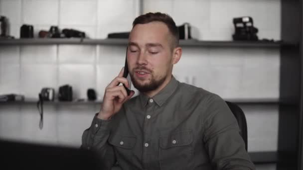 Mladý podnikatel nebo student v košili sedí proti notebooku a mluví se svým obchodním partnerem pomocí chytrého telefonu. Práce na počítači u stolu v kanceláři s přemýšlivým výrazem. — Stock video