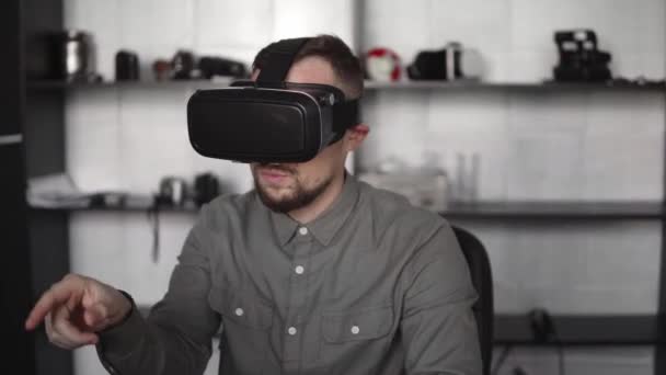 Young bearded man sitting against computer with virtual reality glasses trying to touch something or tap some thing in front of what he seeing. Modern technologies. The concept of future technology. — Stock Video