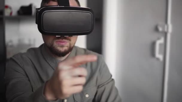 Молодий бородатий чоловік сидить проти комп'ютера з окулярами віртуальної реальності, намагаючись торкнутися чогось або торкнутися чогось перед тим, що він бачить. Сучасні технології. Концепція майбутніх технологій . — стокове відео