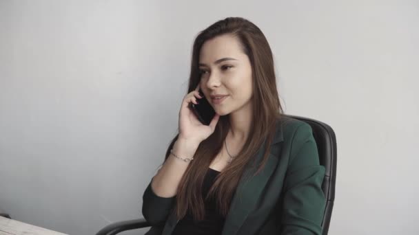 Портрет молодої ділової жінки, яка розмовляє телефоном проти екрану комп'ютера в білому офісі. Жінка-підприємець має дзвінок і роботу з комп'ютером, що сидить за столом. . — стокове відео