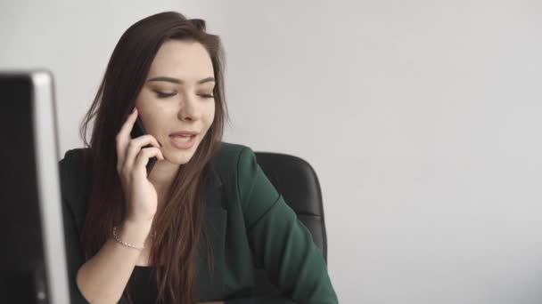 Retrato de una joven mujer de negocios hablando de teléfono contra la pantalla de la computadora en la oficina blanca. Emprendedora que tiene llamada y trabaja con computadora sentada en la mesa.Teléfono llamada de trabajo . — Vídeos de Stock