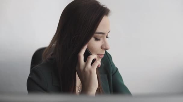 Portrait of young business woman talking phone against computer screen in white office. Female entrepreneur having call and working with computer sitting at table.Telephone work call. — 비디오