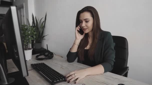 Beyaz ofisteki bilgisayar ekranında telefonla konuşan genç iş kadınının portresi. Kadın girişimci telefonla arıyor ve bilgisayarla çalışıyor. Masada oturuyor. Telefon görüşmesi.. — Stok video