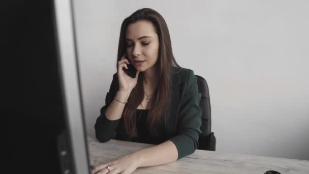 Retrato de una joven mujer de negocios hablando de teléfono contra la pantalla de la computadora en la oficina blanca. Emprendedora que tiene llamada y trabaja con computadora sentada en la mesa.Teléfono llamada de trabajo . — Vídeo de stock