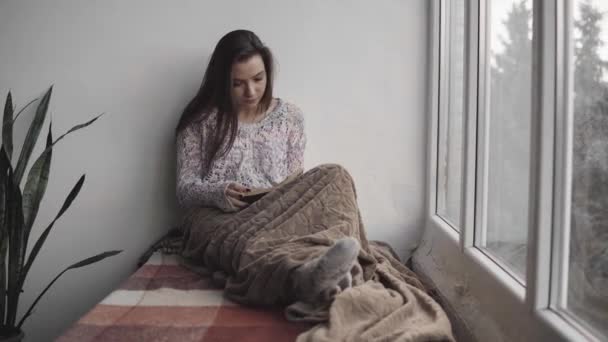 Jolie jeune femme lit le livre assis sur le rebord de la fenêtre dans la maison. Grande fenêtre, plantes vertes . — Video