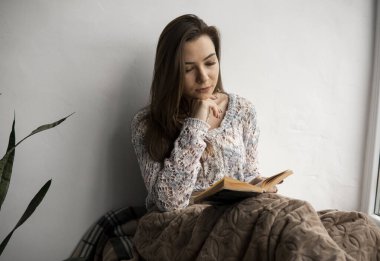 Çekici genç bayan, evin pencere eşiğinde kitap okuyor. Büyük pencere, yeşil bitkiler..