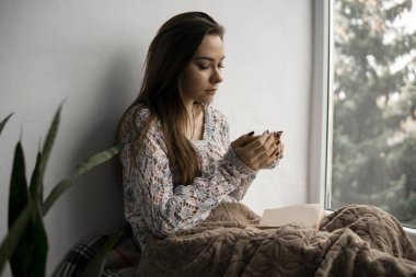 Çekici genç bayan pencere kenarında oturmuş kitap okuyor ve evde çay içiyor. Büyük pencere, yeşil bitkiler..