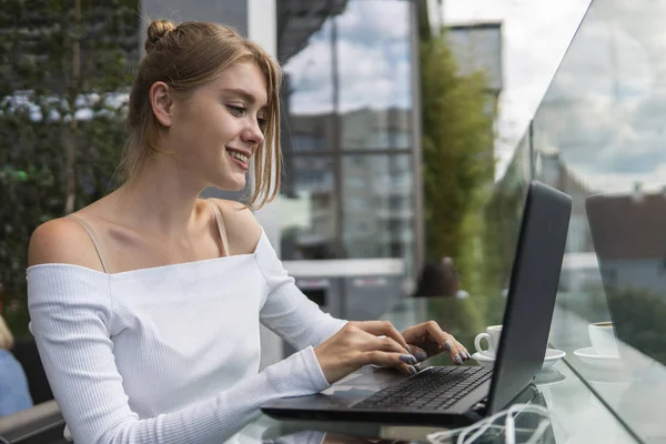Piękna młoda kobieta w białej koszulce pracuje na laptopie i uśmiecha się siedząc na zewnątrz w kawiarni. Młoda kobieta używająca laptopa do pracy. Kobieta freelancer pracuje na laptopie w kawiarni na świeżym powietrzu. — Zdjęcie stockowe