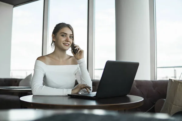 Woman working on laptop at office while talking on phone, backlit warm light. Portrait of young smiling business woman calling her best friend, having break, telling something funny, sitting in cafe. — Stock Photo, Image