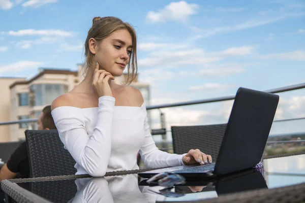Beautiful young woman in white t-shirt is working on laptop and smiling while sitting outdoors in cafe. Young female using laptop for work. Female freelancer working on laptop in an outdoor cafe. — Stock Photo, Image
