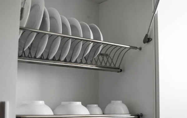 Dish drying metal rack with big nice white clean plates. Traditional comfortable kitchen. Open white dish draining closet with wet dishes of glass and ceramic, plates, bowls drying inside on rack.