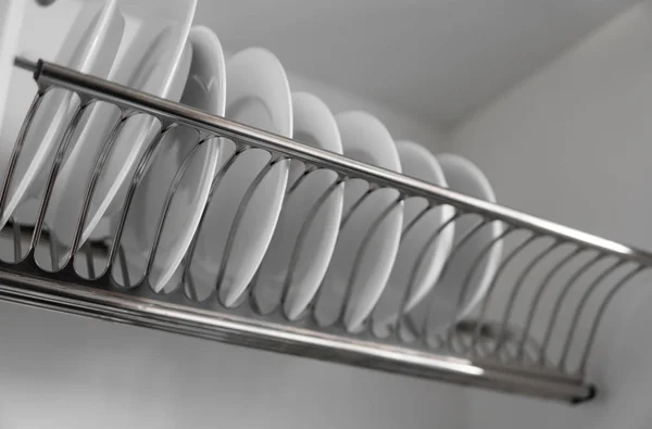 Dish drying metal rack with big nice white clean plates. Traditional comfortable kitchen. Open white dish draining closet with wet dishes of glass and ceramic, plates, bowls drying inside on rack.