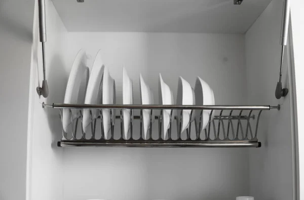 Dish drying metal rack with big nice white clean plates. Traditional comfortable kitchen. Open white dish draining closet with wet dishes of glass and ceramic, plates, bowls drying inside on rack. — Stock Photo, Image