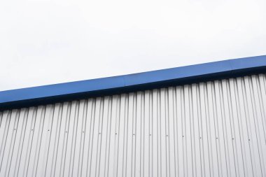 Mavi metal köşeli bir binanın metal kıvrımlı levhaları. Fabrikaların ve sanayi binalarının arka planında bulutlu gökyüzüne karşı beyaz alüminyum metal kıvrımlı çatı veya duvar levhaları.