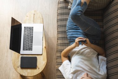 Bir kadın kanepede dizüstü bilgisayar ve masada kitap okurken akıllı telefon kullanır. Çevrimiçi çalışma, serbest çalışma. Serbest çalışan ya da serbest çalışan bir kız telefonunu kullan ve işten izin al..