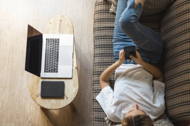 Bir kadın kanepede dizüstü bilgisayar ve masada kitap okurken akıllı telefon kullanır. Çevrimiçi çalışma, serbest çalışma. Serbest çalışan ya da serbest çalışan bir kız telefonunu kullan ve işten izin al..