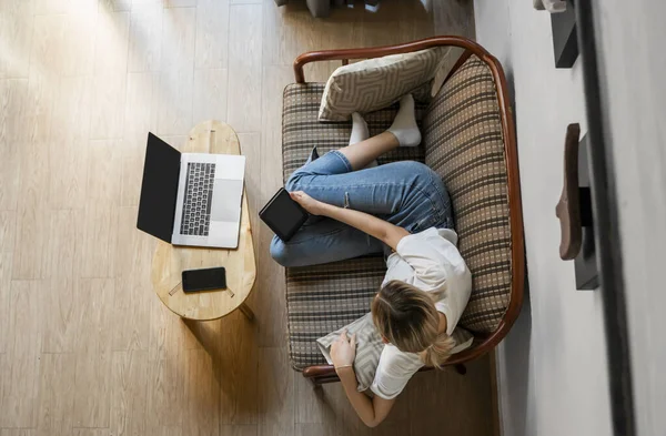Γυναίκα είναι ξαπλωμένη σε έναν καναπέ και χρησιμοποιώντας ένα e-reader ενώ ξεκουράζεται από την εργασία με ένα φορητό υπολογιστή. Μελέτη και εργασία σε απευθείας σύνδεση, ελεύθερος επαγγελματίας. Το κορίτσι δουλεύει με το σημειωματάριό της ξαπλωμένο στον καναπέ.. — Φωτογραφία Αρχείου