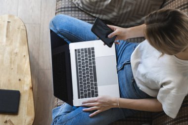 Dizüstü bilgisayarı olan bir kadın kanepede oturuyor ve akıllı telefon kullanıyor. İnternette çalış ve çalış, serbest çalış. Serbest meslek sahibi bir kız defteriyle çalışıyor. Masasında telefon ve okuyucusuyla bir kanepede oturuyor..
