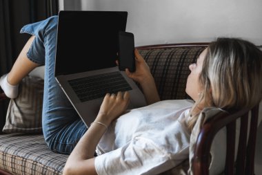 Dizüstü bilgisayarı olan bir kadın kanepede uzanıyor ve akıllı telefon kullanıyor. İnternette çalış ve çalış, serbest çalış. Defteriyle çalışan serbest çalışan bir kız elinde telefonuyla kanepede yatıyor..