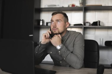 Genç işadamı veya öğrenci bir gömlek bilgisayar monitörü karşı oturan ve akıllı telefon ile iş ortağı ile konuşuyor. Düşünceli bir ifade ile ofiste bir masada bir pc üzerinde çalışma.