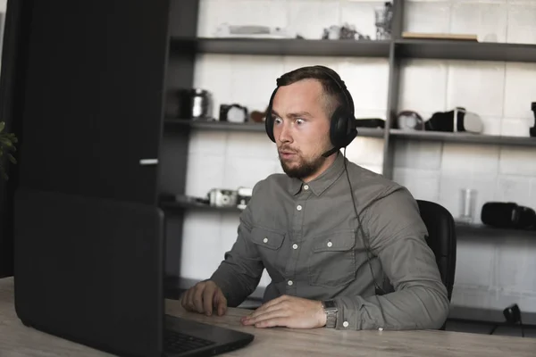 Μπερδεμένος ή θυμωμένος επιχειρηματίας ή μαθητής σε ένα πουκάμισο που κάθεται ενάντια στην οθόνη του υπολογιστή στα ακουστικά και μιλώντας με κάποιον μέσω Διαδικτύου. Εργασία σε έναν υπολογιστή σε ένα τραπέζι στο γραφείο. — Φωτογραφία Αρχείου