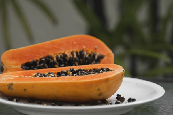 De helft van de rijpe papaya met zaad op een wit bord en met een groene plant op de achtergrond. Snijdt zoete papaja. Halve papaja 's. Gezonde exotische vruchten. Vegetarisch voedsel. — Stockfoto