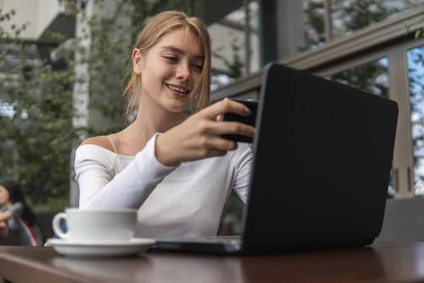 Modern business woman is reading incoming sms message on smartphone or watching broadcasting online on modern mobile phone while sitting in caffe with a laptop. Female student sitting in coffee. — Stock Photo, Image