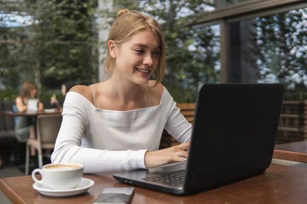 Hermosa mujer joven en camiseta blanca está trabajando en el ordenador portátil y sonriendo mientras se sienta al aire libre en la cafetería. Mujer joven usando el portátil para el trabajo. Freelancer femenina trabajando en un portátil en una cafetería al aire libre . — Foto de Stock