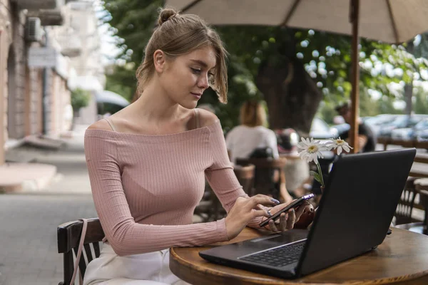 Cheerful blond woman blogger is reading incoming sms message on smartphone connected to free wifi in cafe. Positive young student watching broadcasting online on modern mobile phone. — Stock Photo, Image