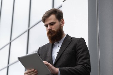Modern genç sakallı iş adamı dijital tabletle çalışıyor. Genç hippi işadamı tableti açık havada tutuyor. Bir ofis binasının önünde dururken bir tabletle çevrimiçi çalışmak.