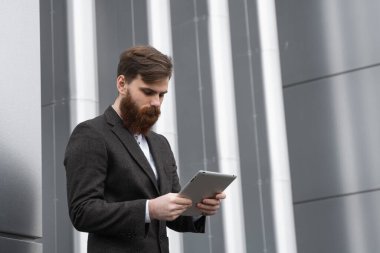 Modern genç sakallı iş adamı dijital tabletle çalışıyor. Genç hippi işadamı tableti açık havada tutuyor. Bir ofis binasının önünde dururken bir tabletle çevrimiçi çalışmak.