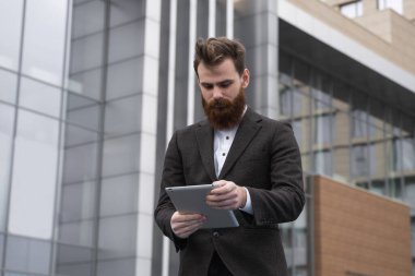 Modern genç sakallı iş adamı dijital tabletle çalışıyor. Genç hippi işadamı tableti açık havada tutuyor. Bir ofis binasının önünde dururken bir tabletle çevrimiçi çalışmak.