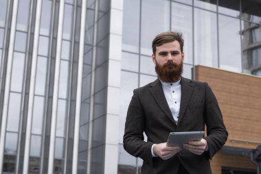 Modern genç sakallı iş adamı elinde dijital tabletle ayakta duruyor. Genç hippi işadamı tableti açık havada tutuyor. Bir ofis binasının önünde dururken bir tabletle çevrimiçi çalışmak.