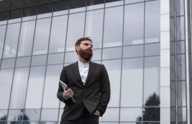 Modern genç sakallı iş adamı elinde dijital tabletle ayakta duruyor. Genç hippi işadamı tableti açık havada tutuyor. Bir ofis binasının önünde dururken bir tabletle çevrimiçi çalışmak.