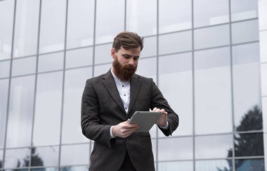 Modern genç sakallı iş adamı dijital tabletle çalışıyor. Genç hippi işadamı tableti açık havada tutuyor. Bir ofis binasının önünde dururken bir tabletle çevrimiçi çalışmak.