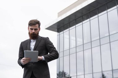 Modern genç sakallı iş adamı dijital tabletle çalışıyor. Genç hippi işadamı tableti açık havada tutuyor. Bir ofis binasının önünde dururken bir tabletle çevrimiçi çalışmak.