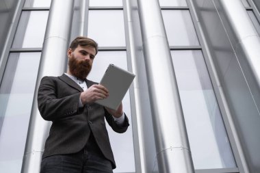 Modern genç sakallı iş adamı dijital tabletle çalışıyor. Genç hippi işadamı tableti açık havada tutuyor. Bir ofis binasının önünde dururken bir tabletle çevrimiçi çalışmak.