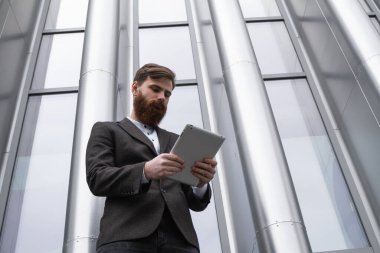 Modern genç sakallı iş adamı dijital tabletle çalışıyor. Genç hippi işadamı tableti açık havada tutuyor. Bir ofis binasının önünde dururken bir tabletle çevrimiçi çalışmak.