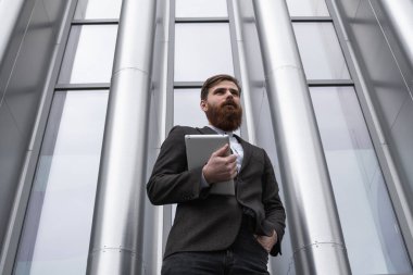 Modern genç sakallı iş adamı elinde dijital tabletle ayakta duruyor. Genç hippi işadamı tableti açık havada tutuyor. Bir ofis binasının önünde dururken bir tabletle çevrimiçi çalışmak.