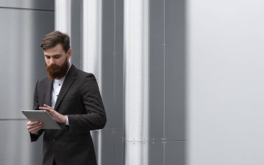 Modern genç sakallı iş adamı dijital tabletle çalışıyor. Genç hippi işadamı tableti açık havada tutuyor. Bir ofis binasının önünde dururken bir tabletle çevrimiçi çalışmak.