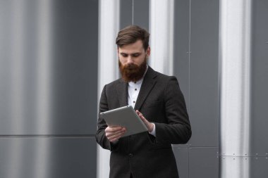 Modern genç sakallı iş adamı dijital tabletle çalışıyor. Genç hippi işadamı tableti açık havada tutuyor. Bir ofis binasının önünde dururken bir tabletle çevrimiçi çalışmak.