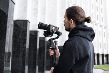 3 eksenli titreşimli dengeleyicide profesyonel kamera tutan genç profesyonel kameraman. Pro ekipmanlar titremeden yüksek kaliteli video çekmeye yardımcı olur. Siyah kapüşonlu bir kameraman video çekiyor..