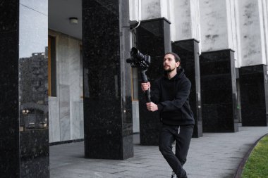 3 eksenli titreşimli dengeleyicide profesyonel kamera tutan genç profesyonel kameraman. Pro ekipmanlar titremeden yüksek kaliteli video çekmeye yardımcı olur. Siyah kapüşonlu bir kameraman video çekiyor..