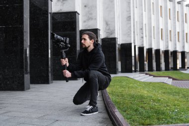 3 eksenli titreşimli dengeleyicide profesyonel kamera tutan genç profesyonel kameraman. Pro ekipmanlar titremeden yüksek kaliteli video çekmeye yardımcı olur. Siyah kapüşonlu bir kameraman video çekiyor..