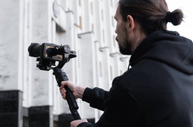3 eksenli titreşimli dengeleyicide profesyonel kamera tutan genç profesyonel kameraman. Pro ekipmanlar titremeden yüksek kaliteli video çekmeye yardımcı olur. Siyah kapüşonlu bir kameraman video çekiyor..
