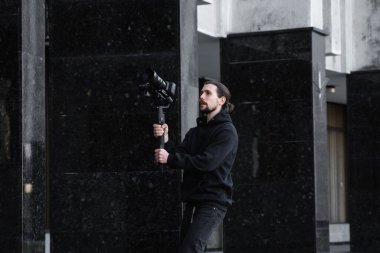 3 eksenli titreşimli dengeleyicide profesyonel kamera tutan genç profesyonel kameraman. Pro ekipmanlar titremeden yüksek kaliteli video çekmeye yardımcı olur. Siyah kapüşonlu bir kameraman video çekiyor..